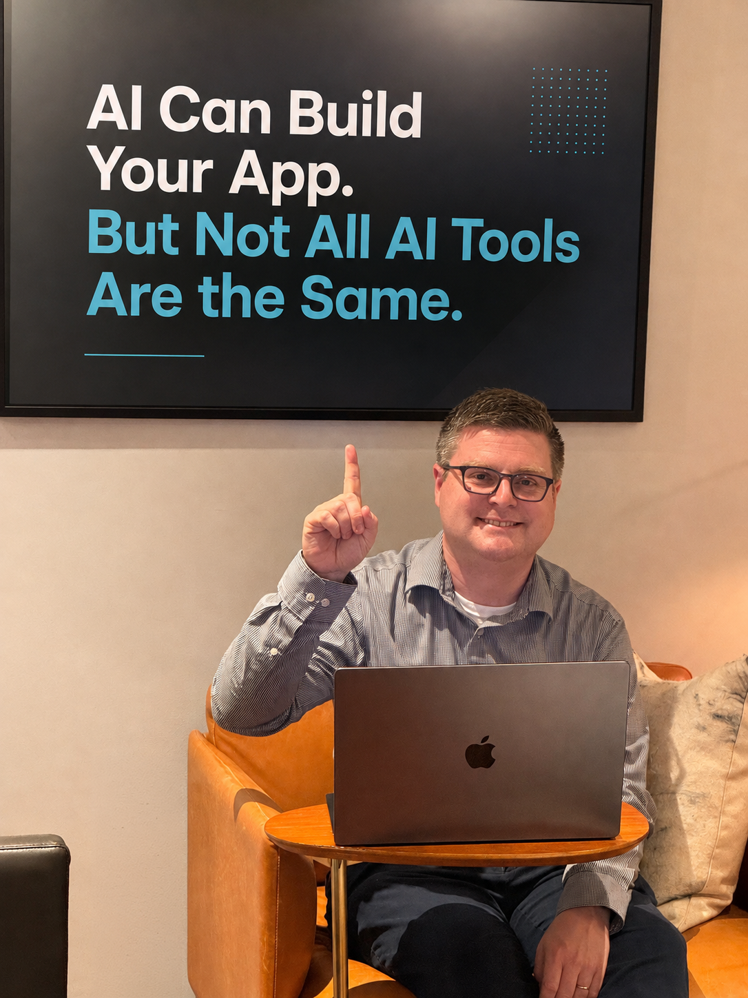 ai-can-build-your-app-not-all-tools-are-the-same Damon Schopen sitting with a laptop in a modern office, pointing to a framed poster that reads, "AI Can Build Your App. But Not All AI Tools Are the Same."